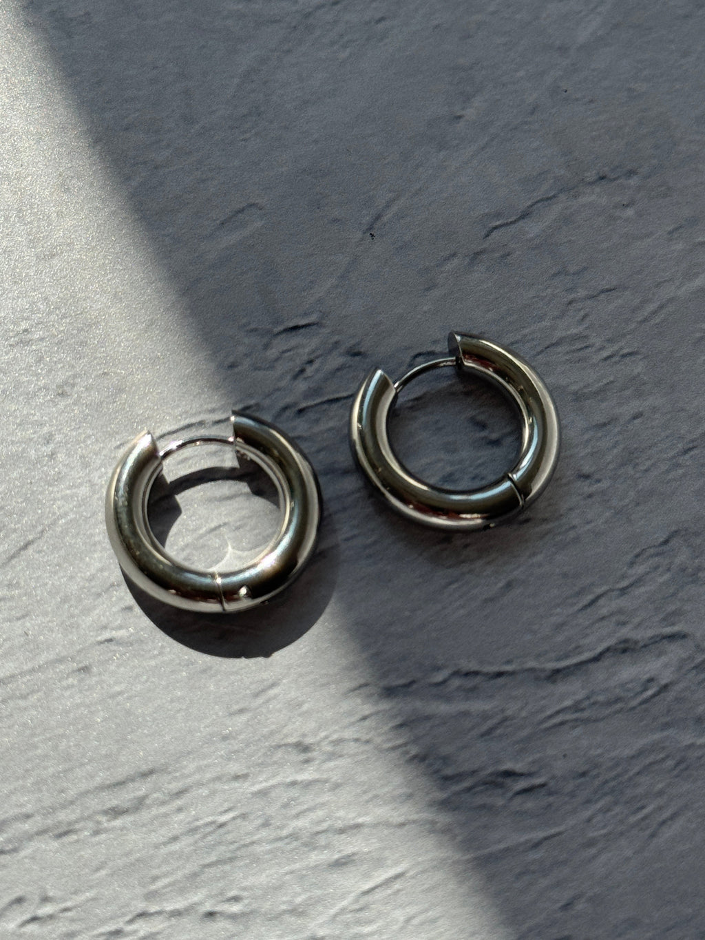 Two silver-toned metal hoop earrings with a split-ring design, resting on a textured gray surface under direct sunlight.