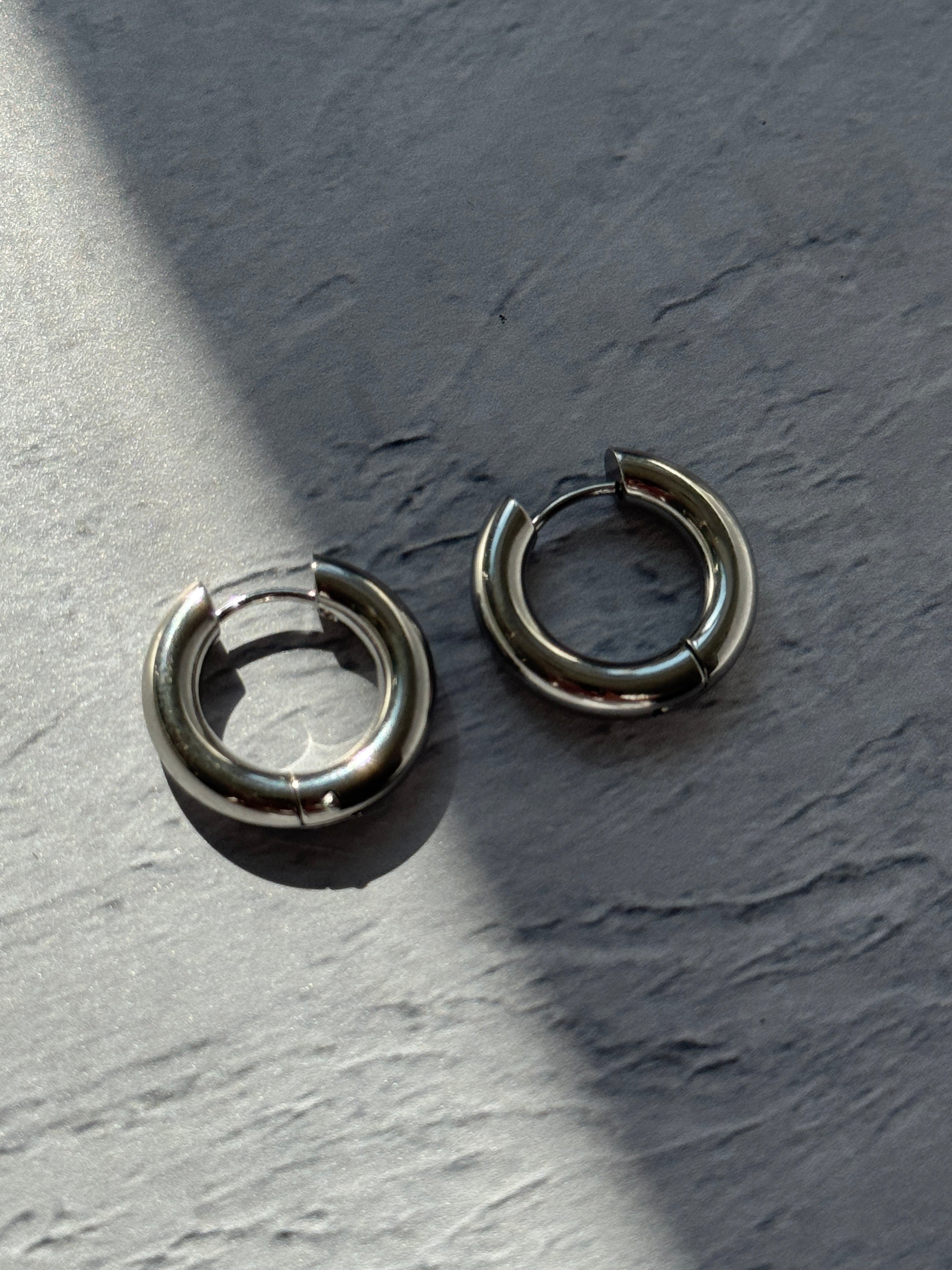 Two silver-toned metal hoop earrings with a split-ring design, resting on a textured gray surface under direct sunlight.