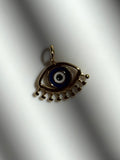 A gold-toned evil eye pendant charm with a blue, white, and black enamel center, resting on a white textured surface with soft shadows.