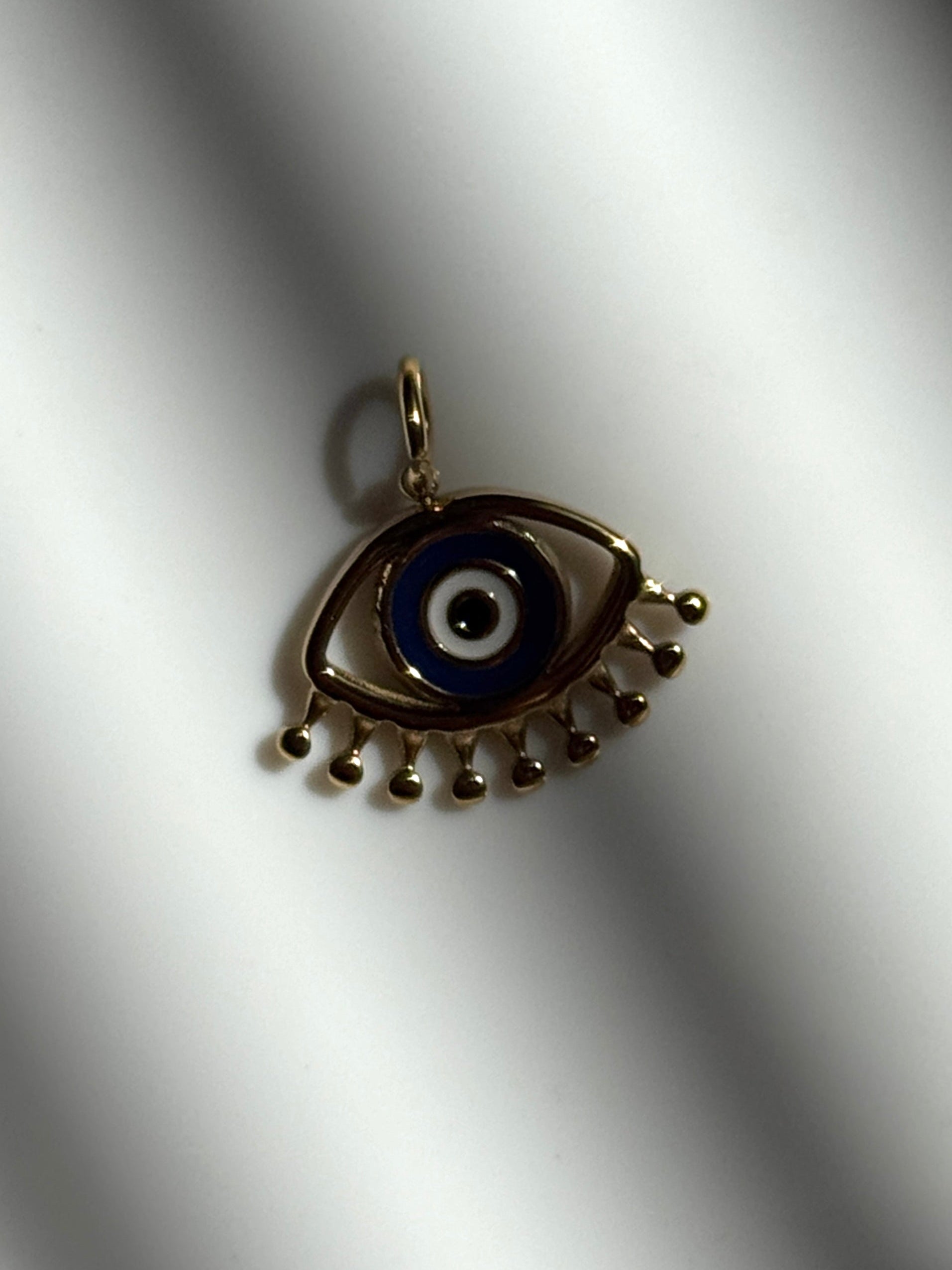 A gold-toned evil eye pendant charm with a blue, white, and black enamel center, resting on a white textured surface with soft shadows.