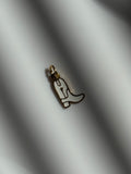 A small cowboy boot charm with a gold-tone outline and white enamel fill, resting on a white surface with soft diagonal shadows.