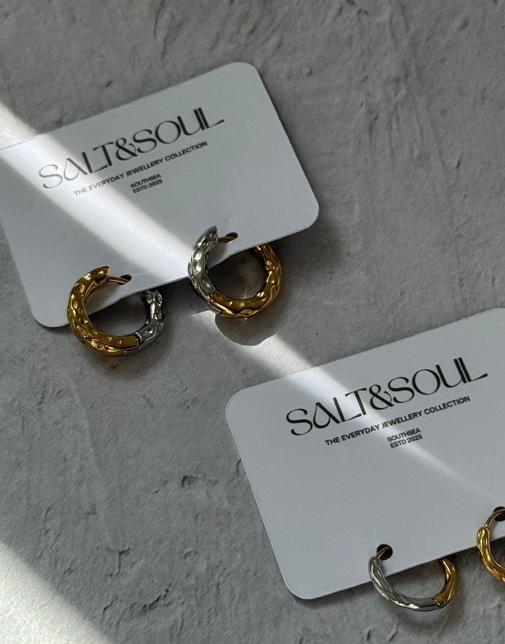 Two pairs of small hoop earrings displayed on white brand cards against a textured light gray surface. The earrings are mixed gold and silver tones, and the cards are printed with the brand name SALT&SOUL.