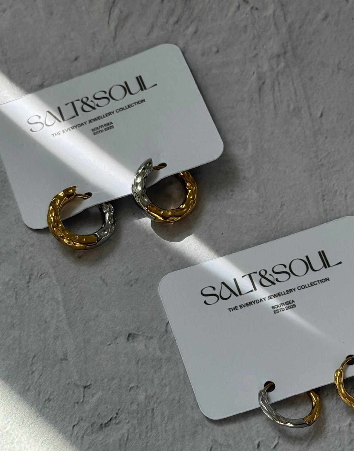 Two pairs of small hoop earrings displayed on white brand cards against a textured light gray surface. The earrings are mixed gold and silver tones, and the cards are printed with the brand name SALT&SOUL.
