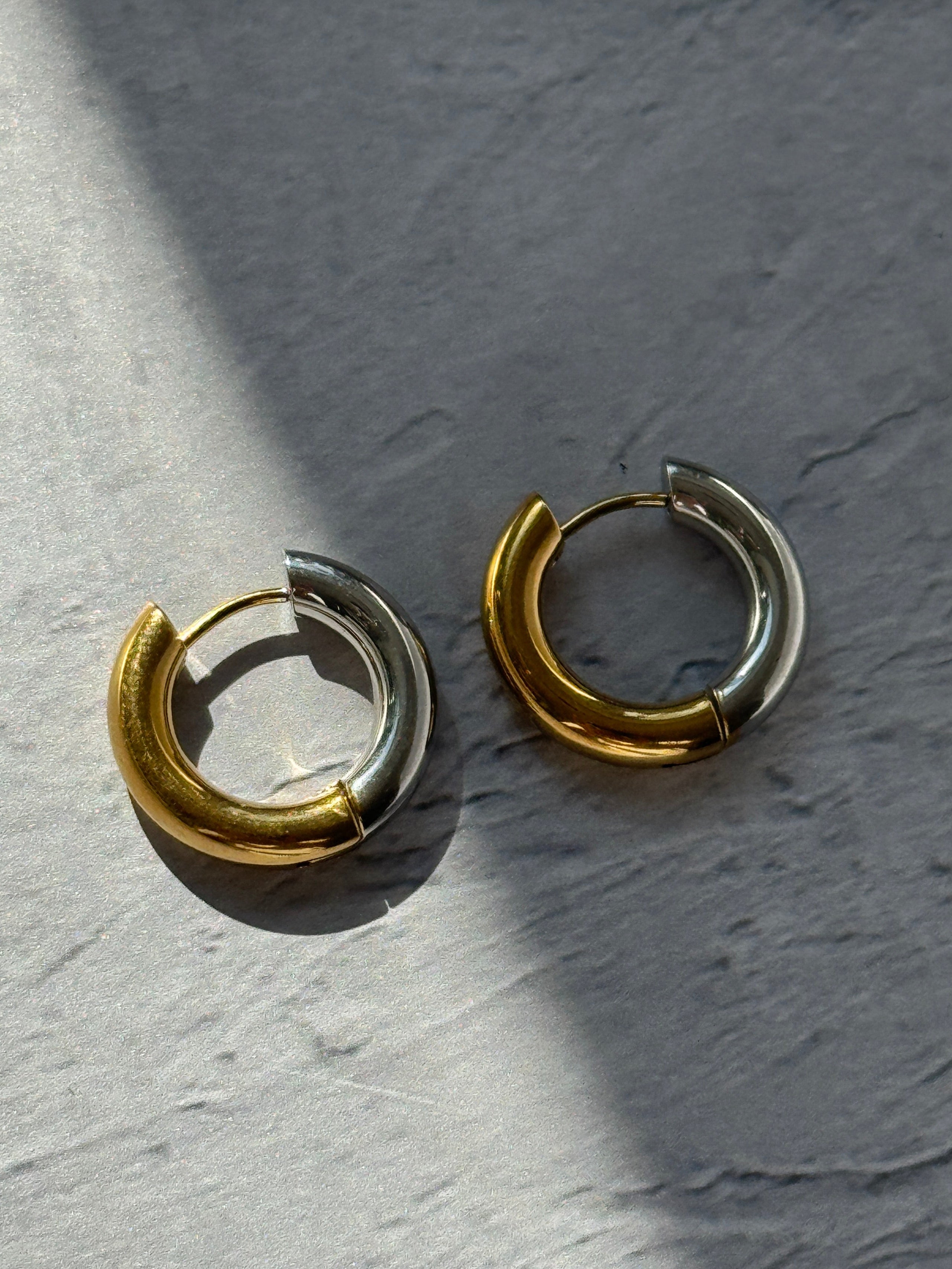 A pair of small split hoop earrings with alternating polished gold and silver halves, resting on a textured gray surface under direct sunlight.