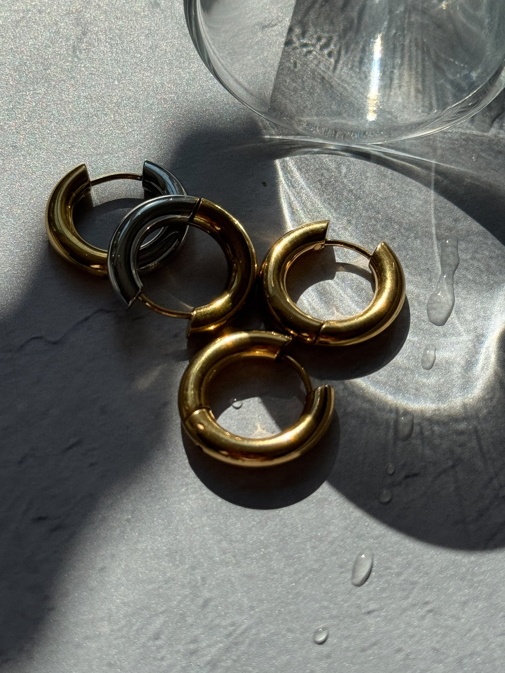 Four chunky split hoop earrings in polished gold and matte silver, arranged on a textured gray surface with water droplets and a glass bowl in the background.