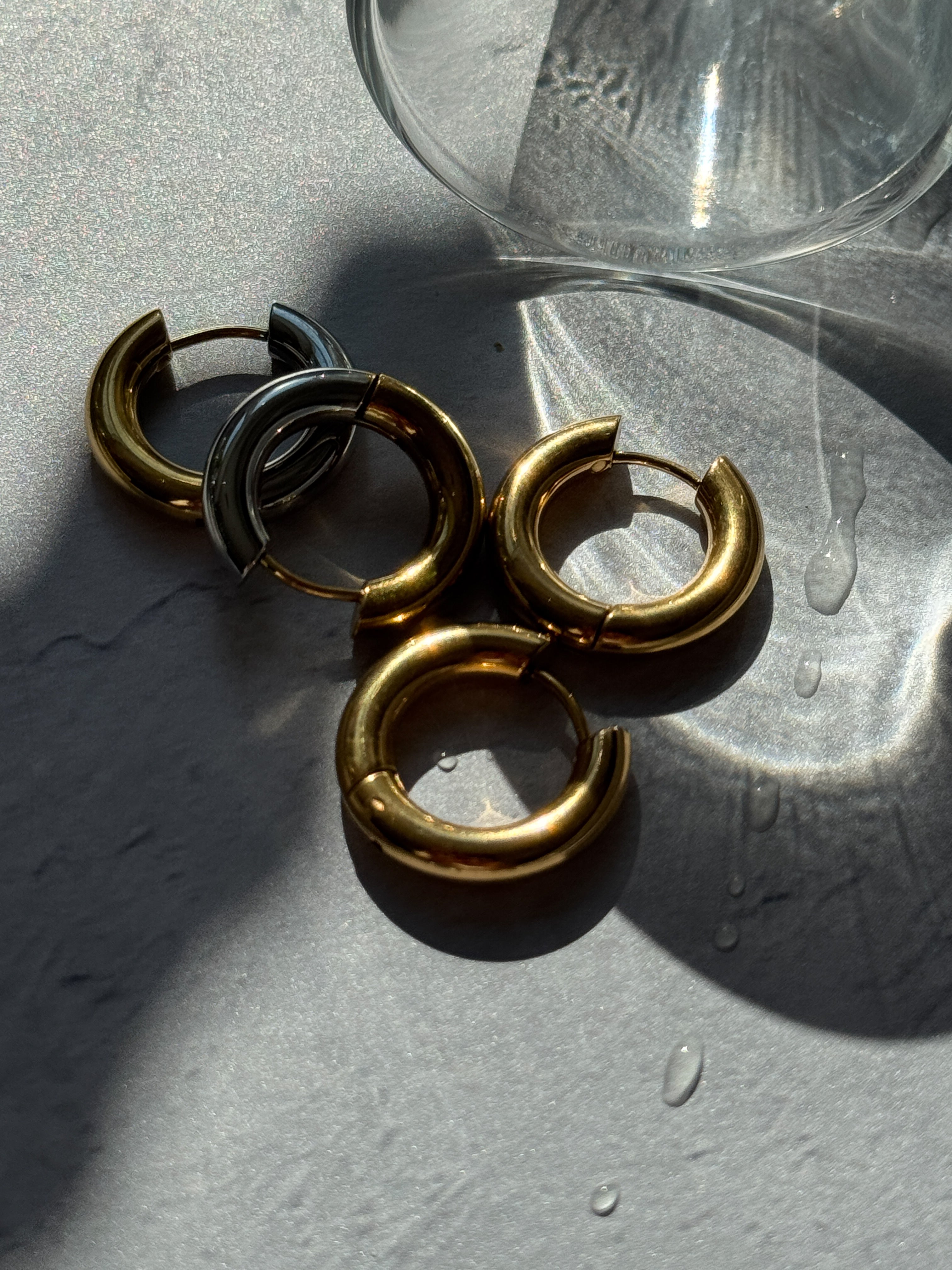 Four chunky split hoop earrings in polished gold and matte silver, arranged on a textured gray surface with water droplets and a glass bowl in the background.