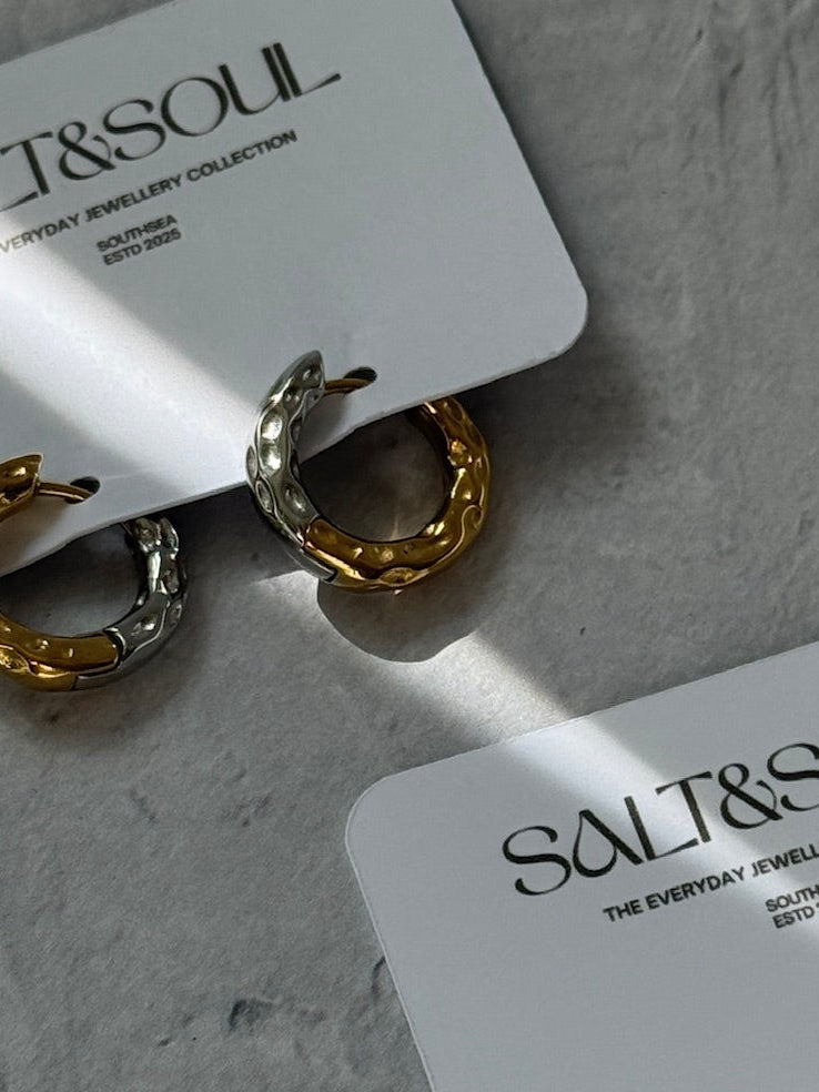 Two pairs of small hoop earrings displayed on white brand cards against a textured light gray surface. The earrings are mixed gold and silver tones, and the cards are printed with the brand name SALT&SOUL.