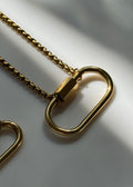 Gold chain necklace with a large open oval ring pendant, photographed on a light gray surface with soft shadows.