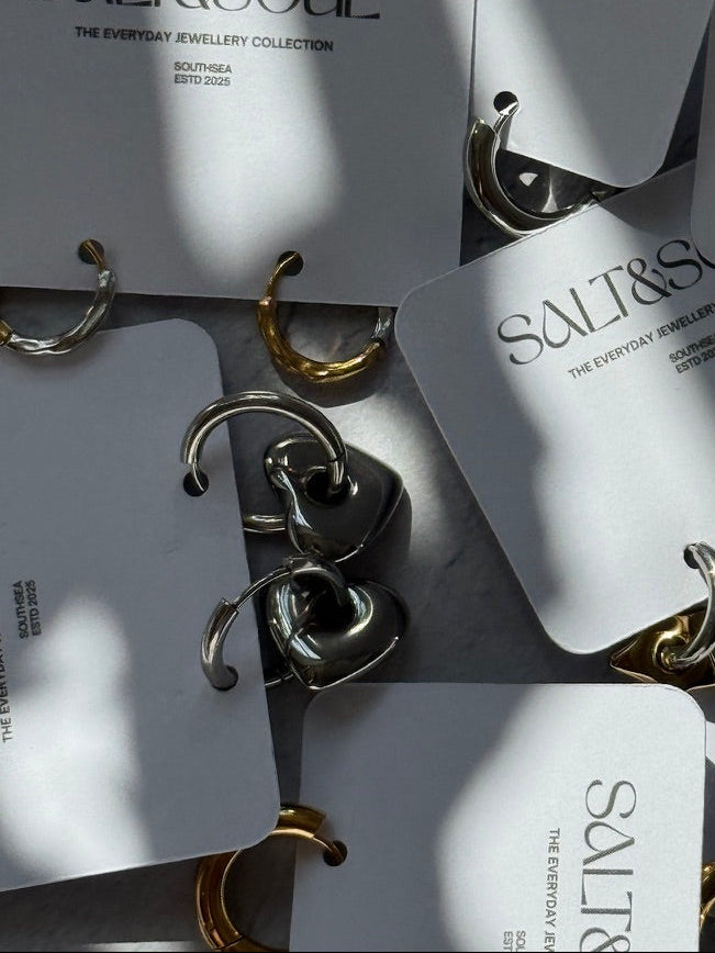 Several small hoop earrings attached to white retail cards printed with the brand name SALT&SOUL. The earrings are shown in both silver and gold finishes, arranged on a light surface with strong natural shadows.