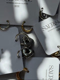Several small hoop earrings attached to white retail cards printed with the brand name SALT&SOUL. The earrings are shown in both silver and gold finishes, arranged on a light surface with strong natural shadows.